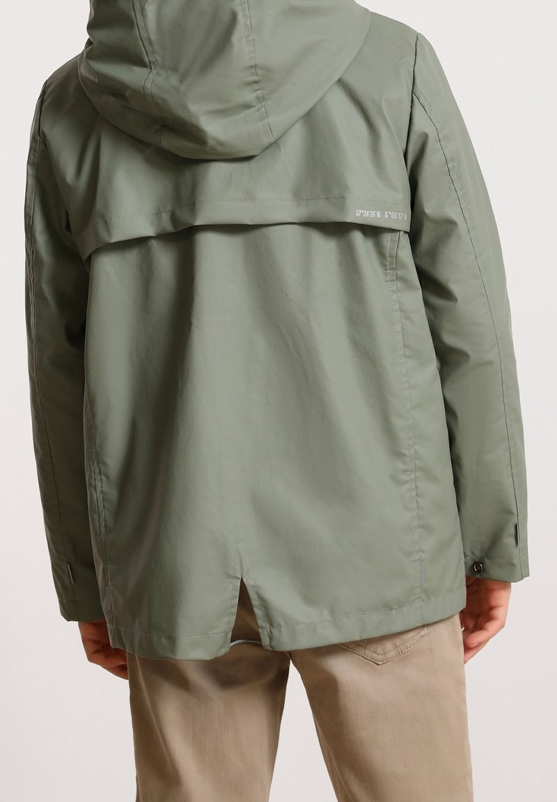 Personne portant une veste à capuche vert olive clair avec un dos ventilé et un petit détail blanc de coordonnées, associée à un pantalon beige.
