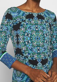 Långärmad klänning med svart bakgrund och blå och gröna paisleymönster, båtknäppning och mönstrade ärmslut med gröna accenter.