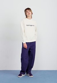 Creme sweatshirt med "carhartt" logo, parret med løstsiddende navy blue bukser og mørke sneakers. Modellen står foran en neutral baggrund.