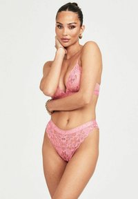 Das Modell posiert in rosa Spitzenunterwäsche mit einem zarten Blumenmuster. Sie stützt eine Hand auf ihr Kinn und präsentiert zarte Schmuckstücke.