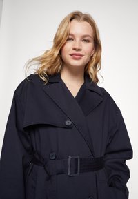 Trench coat blu navy con ampi revers, bottoni doppi e cintura con fibbia. Tessuto morbido con una texture liscia e una forma rilassata.
