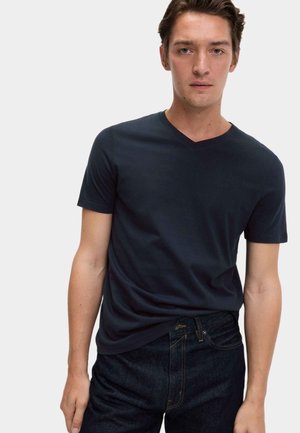 Ung mand iført en ensfarvet mørkeblå V-hals T-shirt og mørke denimjeans, stående mod en ensartet lys baggrund.