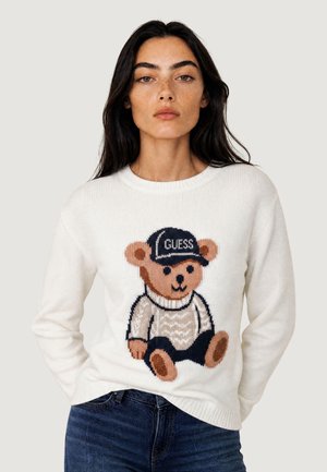 Kobieta w białym swetrze z grafiką misia nosząca czapkę z napisem "GUESS" i ciemnoniebieskie dżinsy, na jednolitym tle.