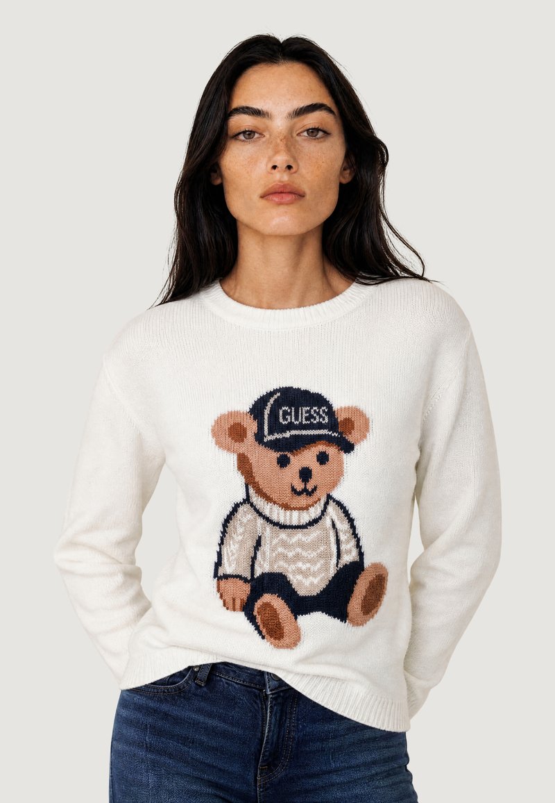 Femme portant un pull blanc avec un motif d'ours en peluche portant une casquette étiquetée "GUESS" et un jean bleu foncé, sur fond uni.