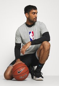 Grå NBA-logo t-shirt over en sort langærmet skjorte, kombineret med sorte shorts med hvide striber. Holder en Nike basketball.