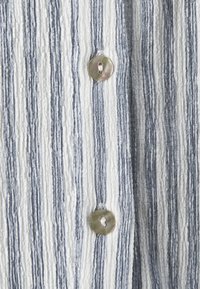 Wit en blauw gestreept stof met verticale strepen en een getextureerde afwerking, met een ronde, iriserende knoop in het midden.