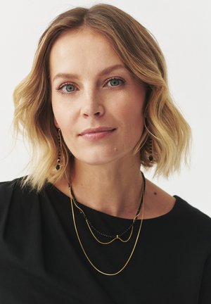 Gold and black layered necklaces worn over a black top. The necklaces feature varying chain lengths and bead accents. Smooth texture.