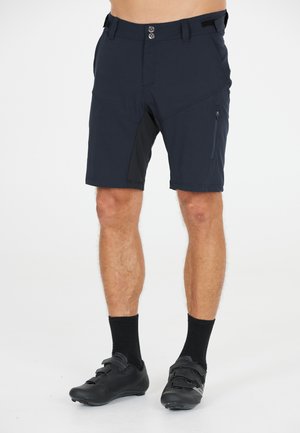Marineblauwe fietsshort met een gestructureerde afwerking, voorzien van twee voorzakken en een aansluitend ontwerp. Gepaarde met zwarte fietsschoenen en sokken.