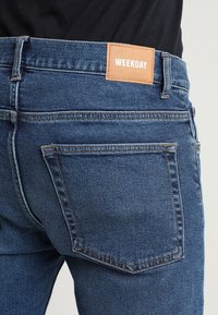 Gros plan sur la poche arrière d'un jean en denim bleu avec une étiquette en cuir marron portant le mot "WEEKDAY" sur une personne portant un haut noir.