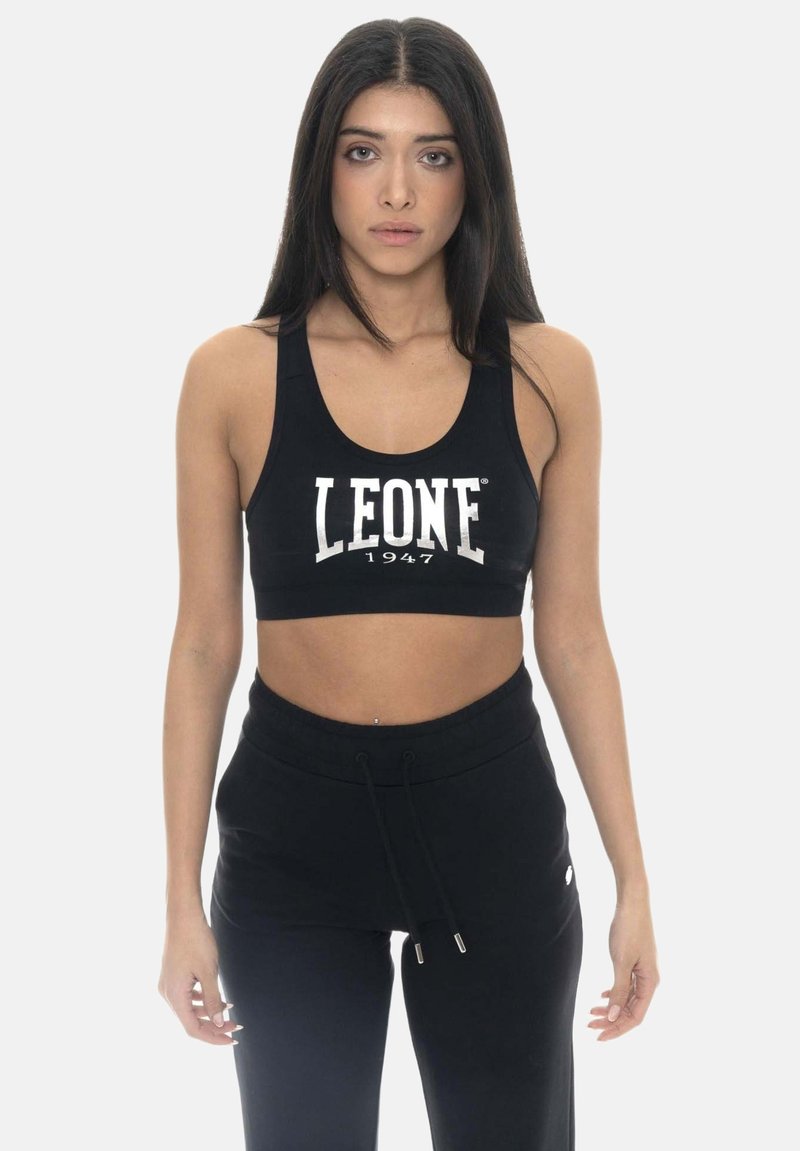 Femme aux longs cheveux foncés portant un soutien-gorge de sport noir avec le texte « "LEONE 1947" » et un pantalon noir à cordon, debout devant un fond uni.