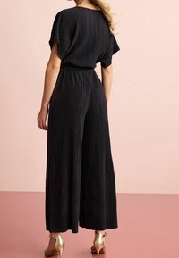 Zwarte, geplooide jumpsuit met wijde benen en korte mouwen. Heeft een versmalde taille. Gepaarde met gouden haksandalen.