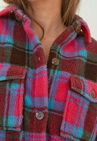 Fleece košile v jasně růžové, modré a hnědé kostkované vzoru. Má zapínání na knoflíky vpředu a náprsní kapsy s kulatými knoflíky. Texturovaný materiál.