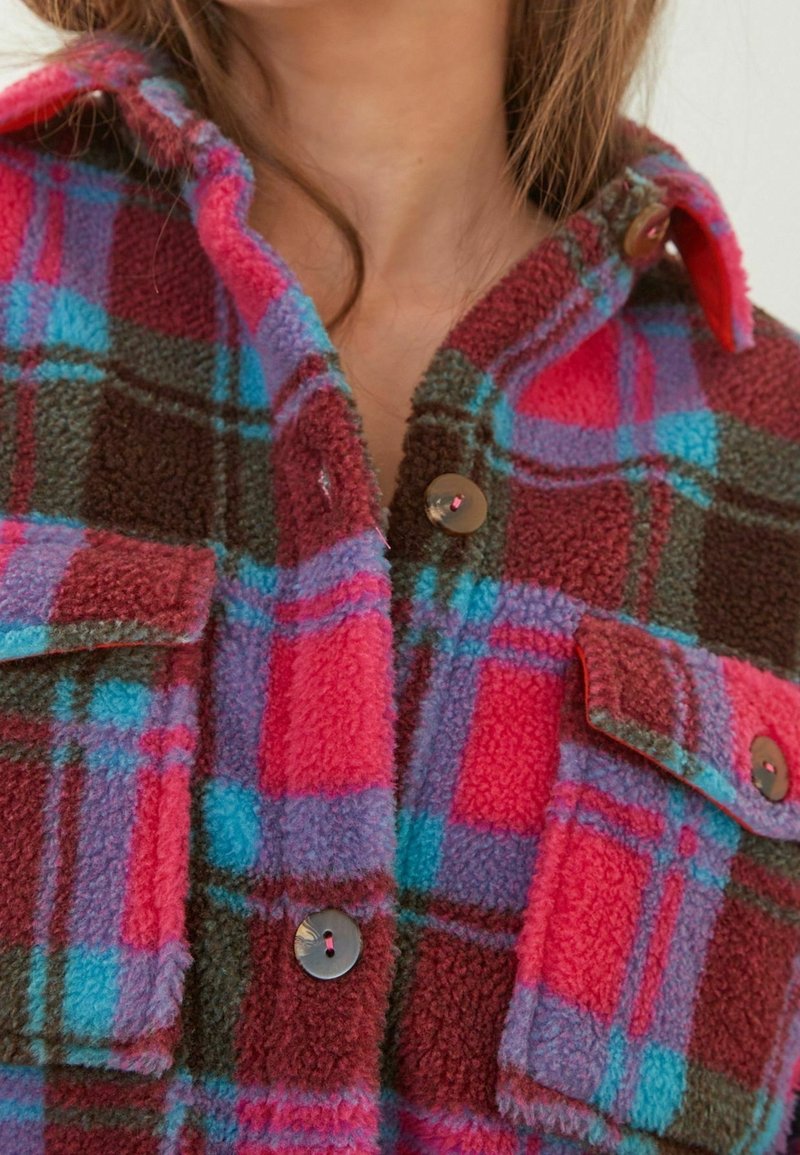 Fleece košile v jasně růžové, modré a hnědé kostkované vzoru. Má zapínání na knoflíky vpředu a náprsní kapsy s kulatými knoflíky. Texturovaný materiál.