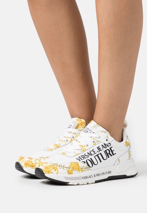 Witte sneakers met gele en zwarte decoratieve patronen, voorzien van het merk "VERSACE JEANS COUTURE" en een geribbelde rubberen zool.