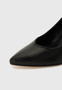 Scarpa con tacco alto in pelle verniciata nera, con punta affusolata e texture a motivo rettile. Il design presenta dei fori sui lati.