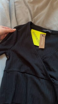 Zwarte, geribbelde wikkeltop van biologisch katoen. Heeft een V-hals en korte mouwen. Het label dat eraan vastzit toont de merknaam en materiaaldetails.