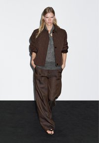 Frau, die auf einem schwarzen Laufsteg geht, trägt eine braune gesteppte Jacke, ein graues Strickoberteil, weite braune Hose und offene Sandalen, Hände in den Taschen, neutrale Miene.