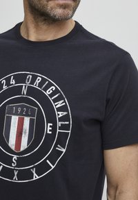 Sötétkék, rövid ujjú pamut póló, kör alakú, fehér grafikával, amelyen pajzs design és a "24 ORIGINAL" valamint a "1924" szöveg szerepel.