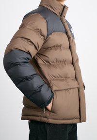 Giacca a ventosa marrone con sezioni nere sulle spalle e sulle braccia, dotata di chiusura con zip e tasche esterne. Il materiale è liscio e imbottito.