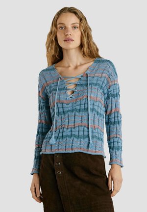 Femme portant un haut à manches longues texturé bleu et rose à rayures avec un décolleté lacé en croix et une jupe boutonnée marron foncé, fond neutre.