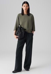 Olijfgroen oversized top met een ronde hals, gecombineerd met losse zwarte pantalon. De figuur houdt een zwarte tas met schouderbanden vast.