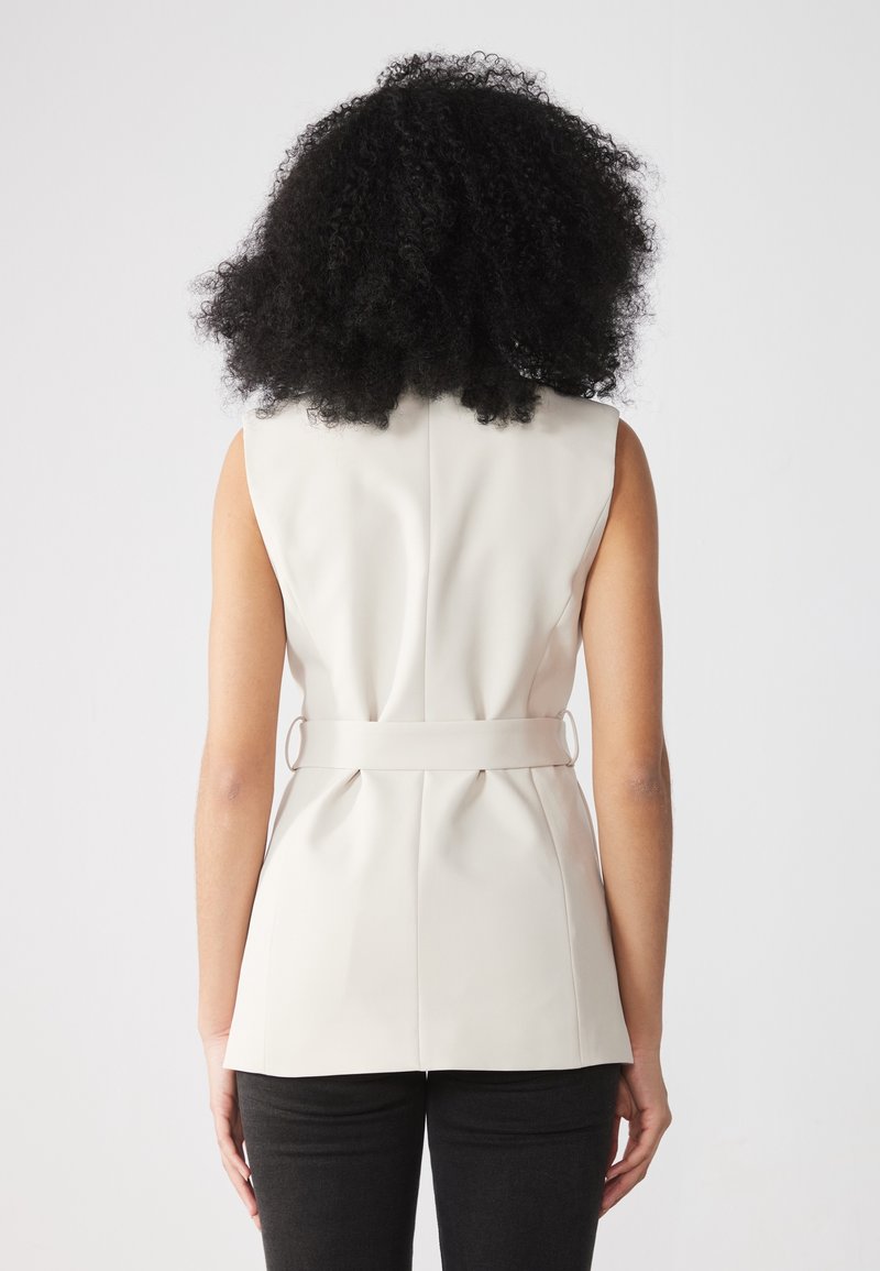 Femme aux cheveux noirs bouclés portant un gilet blanc sans manches ceinturé et un pantalon noir, debout devant un fond blanc uni.