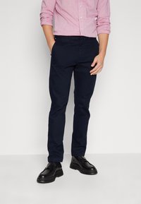 Chino blu navy realizzati in cotone, con una vestibilità slim, tasche laterali e una texture liscia. Indossati con una camicia a fantasia e scarpe nere.