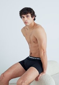 Boxer-briefs noirs avec une taille à logo, portés par un mannequin assis contre un fond clair, mettant en valeur un tissu lisse et un design ajusté.