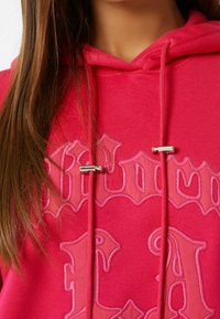 Fuchsiový svetr s vyšitým textem, doplněný stříbrnými akcenty na šňůrkách a měkkou texturou. Dlouhé rukávy, s kapucí.
