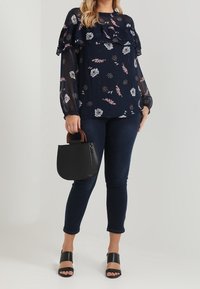 Marineblaue Bluse mit Blumenmuster und geschichteten Rüschen, kombiniert mit dunklen Jeans, schwarzen offenen Absatzschuhen und einer kleinen schwarzen Handtasche mit Holzgriff.