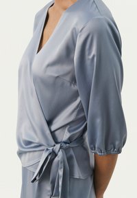 Blusa di satin azzurro chiaro con un design avvolgente, scollo a V profondo, maniche a sbuffo corte e un dettaglio annodato in vita. Tessuto liscio.