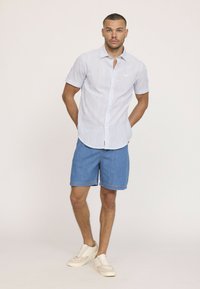 Chemise à manches courtes rayée bleu clair associée à un short en denim. Le mannequin porte des baskets blanches. La chemise dispose d'une fermeture à boutons et d'un ourlet arrondi.