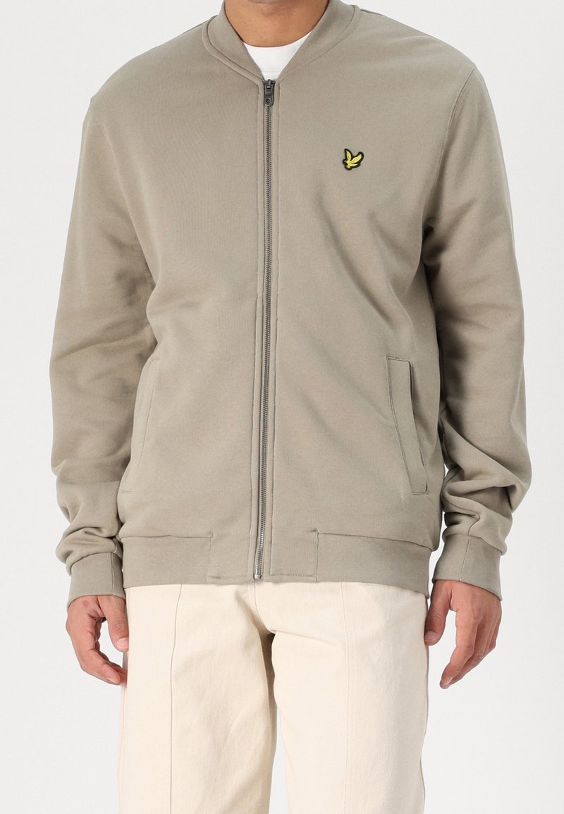 Homme portant un sweat-shirt beige zippé avec un petit logo oiseau jaune sur la poitrine et un pantalon blanc cassé, debout devant un fond uni.