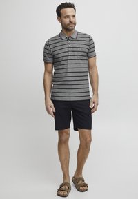Grå og svart stripete polo skjorte med korte ermer, krage, rød kant og brystlomme, kombinert med mørke shorts og brune sandaler.
