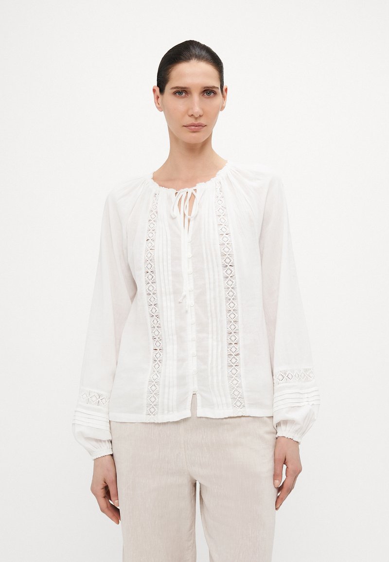 Hvit linbluse med lange pufferm, rund hals med knyting og dekorative blonde detaljer langs fronten og ermene.