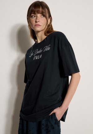 Junge Frau mit braunen Haaren, die ein schwarzes übergroßes T-Shirt mit dem Schriftzug "La Dolce Vita Italia" trägt und vor einem einfarbigen hellen Hintergrund steht.