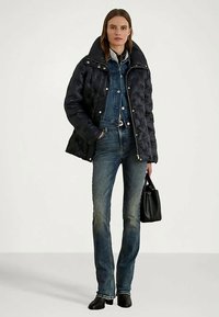 Γυναίκα ντυμένη με ένα μαύρο επενδυμένο μπουφάν πάνω από ένα μπλε τζιν μπουφάν και τζιν, κρατώντας μια μαύρη τσάντα, που στέκεται σε ένα λευκό φόντο.