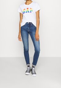 Camiseta blanca con el logo 'GAP' multicolor, combinada con jeans ajustados de color azul oscuro y zapatillas altas negras. Tejido suave y corte casual.
