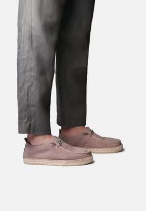 Osoba u tamnosivim skraćenim hlačama i svijetlosmeđim antilop mokasinama s tkanim jutenim potplatom.