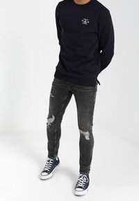 Sudadera de marina con cuello redondo y logo blanco. Jeans ajustados negros desgastados con detalles rasgados. Zapatillas altas azules.
