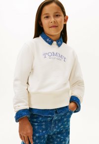 Felpa bianca con logo "TOMMY HILFIGER" in lilla chiaro, abbinata a un colletto in denim blu e pantaloni blu a fantasia con dettagli floreali.