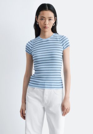 Jeune femme aux longs cheveux noirs portant des boucles d'oreilles en créole, un haut ajusté à manches courtes bleu clair à motifs, et un pantalon blanc, posant devant un fond blanc.