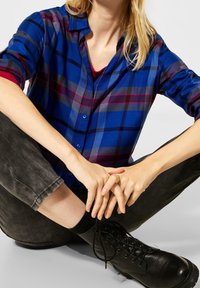 Blue plaid button-up shirt with purple and gray accents, worn over a red top. Black high-ankle boots and gray fitted pants.