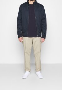 Marinblå bomberjacka med dragkedja, ribbade ärmslut och ståkrage, i kombination med en mörk topp, beiga byxor och vita sneakers.