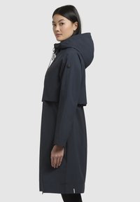 Parka imperméable bleu marine avec un design cropped, capuche, accents à cordon de serrage, manches longues et poches latérales. Texture lisse et style minimaliste.