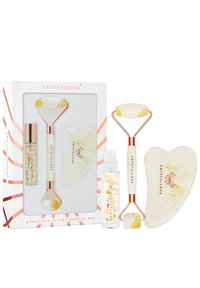 Zestaw do pielęgnacji twarzy z kamieniami szlachetnymi, zawierający wałek z białego i żółtego jadeitu, sercowy guasha oraz szklany flakon z białymi kryształami, zapakowany w pudełku.