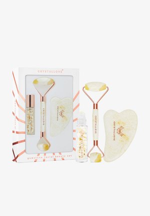 Zestaw do pielęgnacji twarzy z kamieniami szlachetnymi, zawierający wałek z białego i żółtego jadeitu, sercowy guasha oraz szklany flakon z białymi kryształami, zapakowany w pudełku.