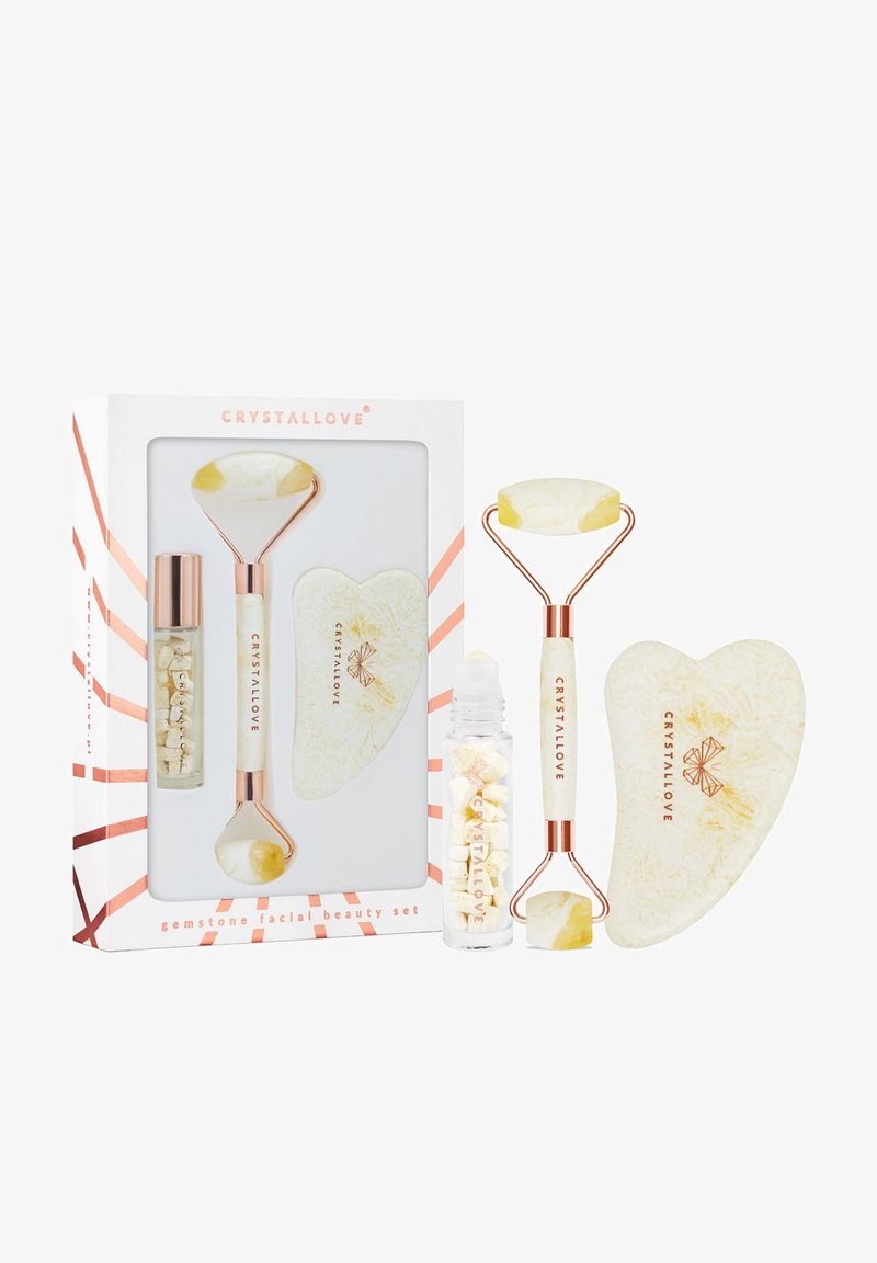 Zestaw do pielęgnacji twarzy z kamieniami szlachetnymi, zawierający wałek z białego i żółtego jadeitu, sercowy guasha oraz szklany flakon z białymi kryształami, zapakowany w pudełku.
