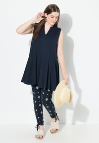Top senza maniche di colore blu navy con design plissettato, abbinato a leggings a fantasia con forme geometriche. Sandali con zeppa di colore chiaro e cappello di paglia.