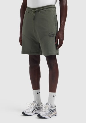 Person trägt olivgrüne Shorts mit Kordelzug, weiße Nike-Socken und silberne Laufschuhe und steht vor einem einfarbigen hellen Hintergrund.